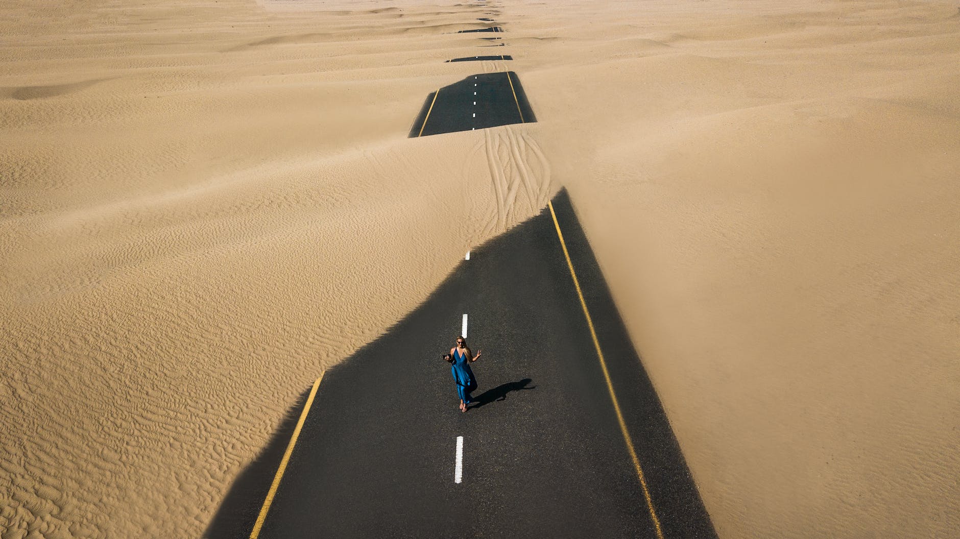 The Laguna Bungle. A highway through a desert is partially covered by sand. A woman in a blue dress stands in the middle of it. 