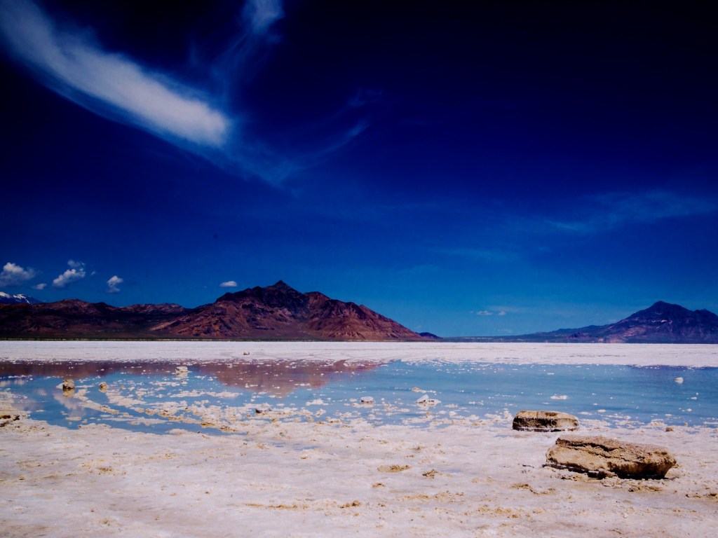 White salt flats surrounding a shallow pool of light blue water. There are brownish, jagged mountains in the background below a deep blue sky.